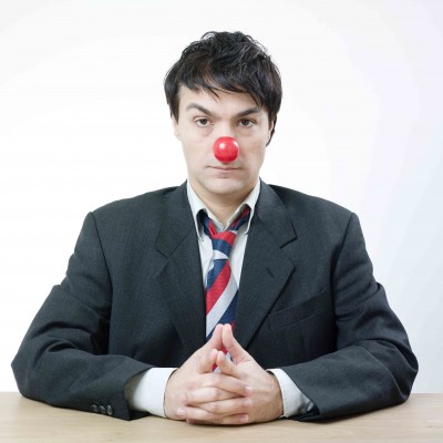 A portrait of a caucasian man wearin suit and necktie and looking serious .Paradoxically, he is wearing a red nose .